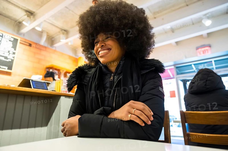 Smiling black woman in a winter jacket chatting happily inside a bustling manhattan coffee shop, enjoying a cozy, relaxed moment of urban comfort and confident contentment