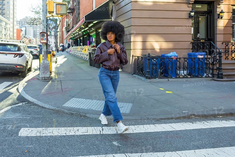 Young woman with afro hair and casual fashion attire confidently walking across a zebra crossing in a vibrant new york city urban street, representing city life and modern style