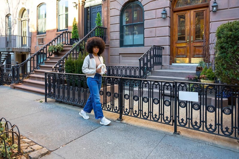 Young woman with afro hair walking down a sidewalk in a historical new york city neighborhood, holding a fashionable accessory and exploring the urban environment