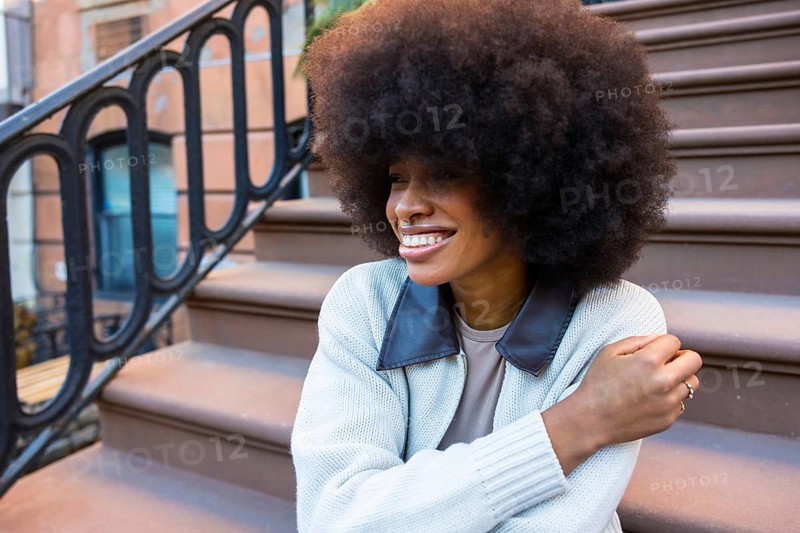 African american woman with a beautiful afro hairstyle sitting and smiling on brownstone steps in manhattan, embodying urban lifestyle, joy, and individual expression
