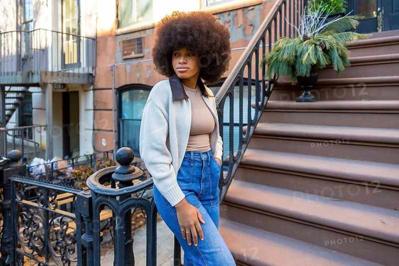 Stylish young woman with afro hair posing confidently on the brownstone steps of an urban building, embodying modern street fashion and a vibrant city lifestyle in harlem, new york