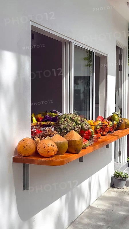Assortment of fresh tropical fruit and young raw coconuts displayed on a wooden shelf outside a building window, offering healthy refreshments in a sunny outdoor setting