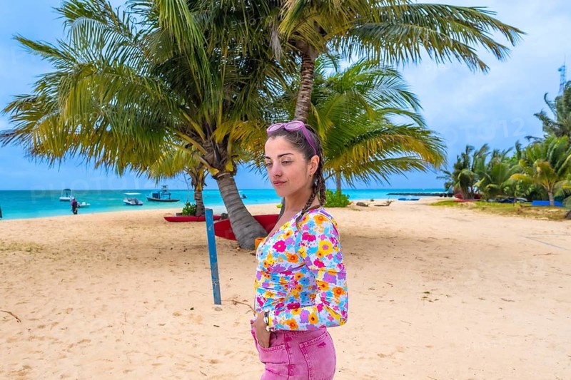 Young woman posing on sandy beach beneath two palm trees, smiling and enjoying a tropical island getaway with clear blue water, boats on the horizon and sunny skies