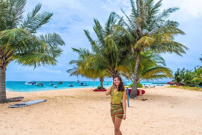 Woman smiling and standing on a beautiful sandy beach fringed with green palm trees and turquoise ocean water with boats in the background, representing travel and leisure