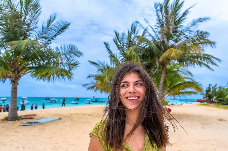 Young woman smiling and looking away, standing on a sandy beach with palm trees and a turquoise ocean bustling with boats in the background, embodying travel, freedom, and relaxation