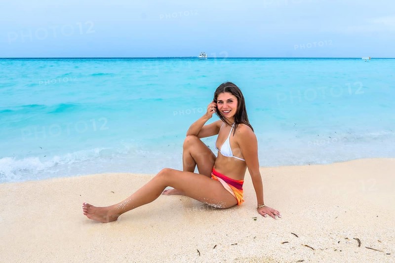 Young woman sitting on white sand beach smiling, enjoying a relaxing tropical getaway with clear turquoise ocean, distant boat and sunny blue sky on an island resort