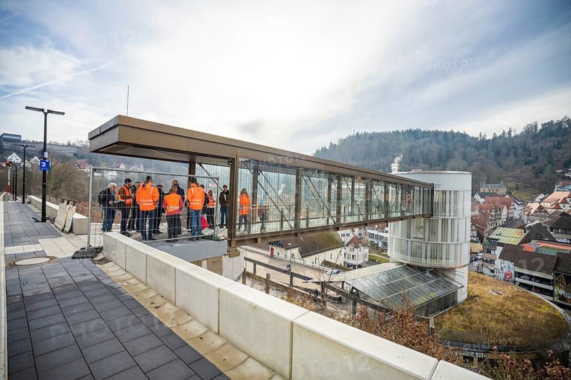 People in orange vests stand on a glass bridge with a view of a city and hills, taking off the Hermann Hessebahn, Calw, Germany