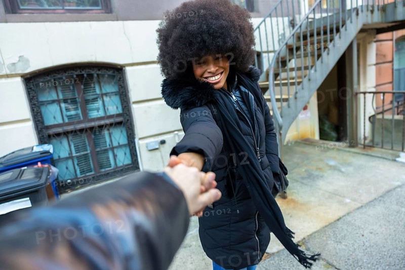 Young black woman smiling, looking back, and holding a hand, guiding through a city street in manhattan with a happy expression and engaging gesture, dressed in a winter jacket