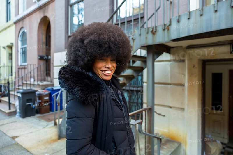 Smiling woman with a large afro hairstyle standing outdoors on a city street, wearing a winter jacket near brownstone buildings and a spiral fire escape, depicting urban lifestyle and natural beauty