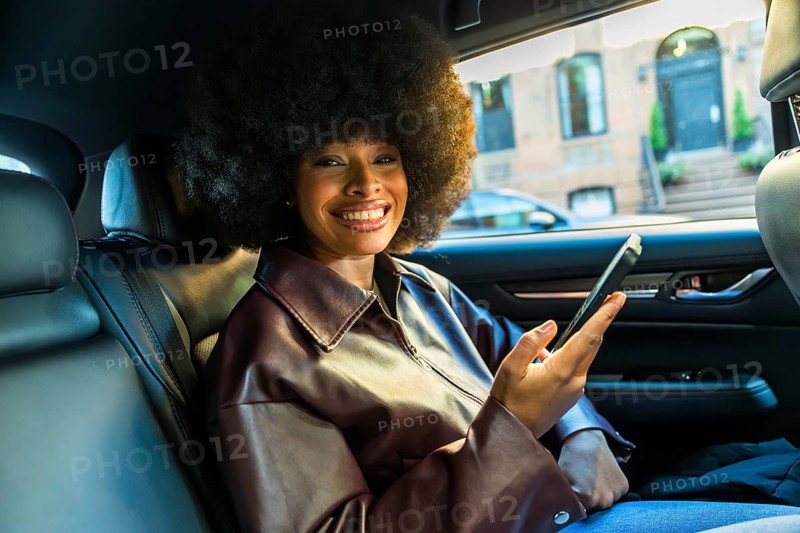 Young black woman with an afro hairstyle smiling at the camera and holding a smartphone while sitting in the backseat of a car, enjoying an urban journey through manhattan