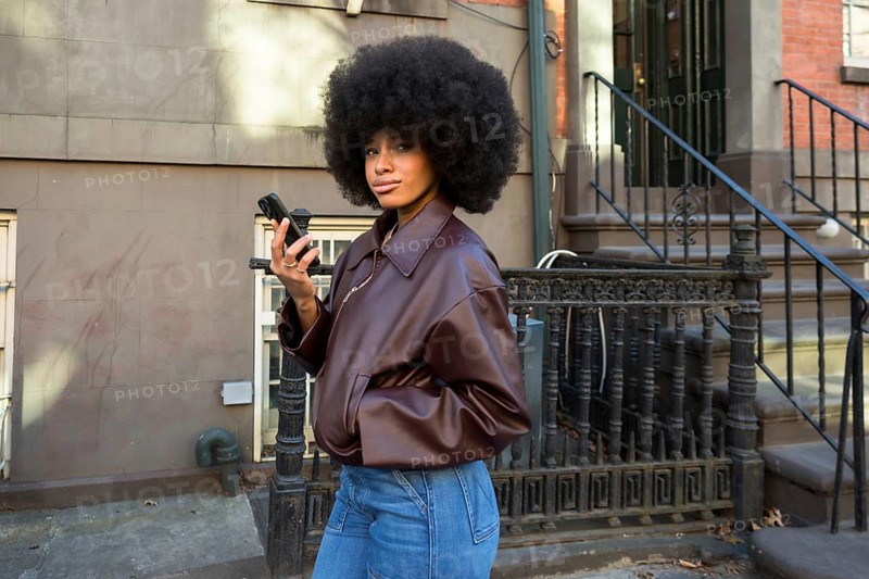 Young woman with afro hair wearing a brown jacket and jeans, looking at the camera while holding a mobile phone, representing urban lifestyle and connectivity in new york city