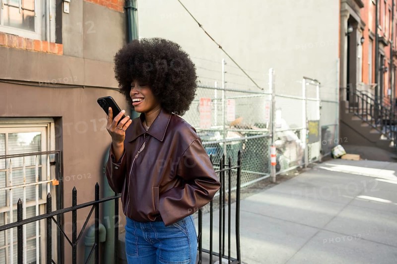 Young african american woman with afro hairstyle laughing while sending an audio message on her smartphone, enjoying urban street life on a sunny day in manhattan