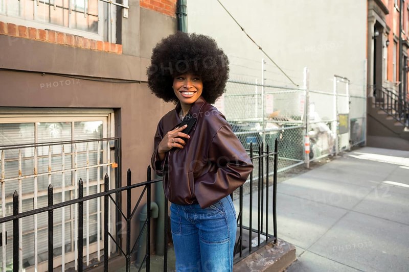 Young african american woman with a large afro hairstyle smiling, wearing a leather jacket and jeans, standing beside a fence on an urban sidewalk in new york city