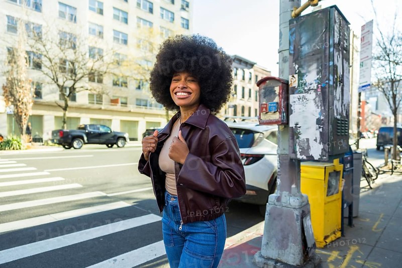 Young african american woman with a big afro hairstyle and smiling face standing on a new york city street, wearing a brown jacket and jeans, representing urban joy and diverse lifestyle