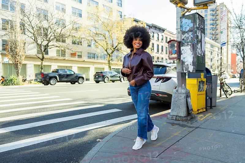 Young black woman smiling confidently, walking briskly on a manhattan city street, wearing a stylish leather jacket and jeans, capturing urban lifestyle and fashion trends