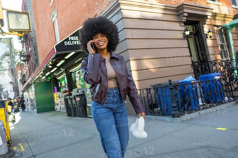 Happy young woman with an afro smiling widely, talking on her mobile phone while walking along a bustling sidewalk in manhattan, united states, enjoying urban life