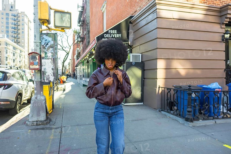 Young black woman with an afro hairstyle adjusting her brown leather jacket while standing on a manhattan street sidewalk, showcasing urban fashion in a vibrant city environment