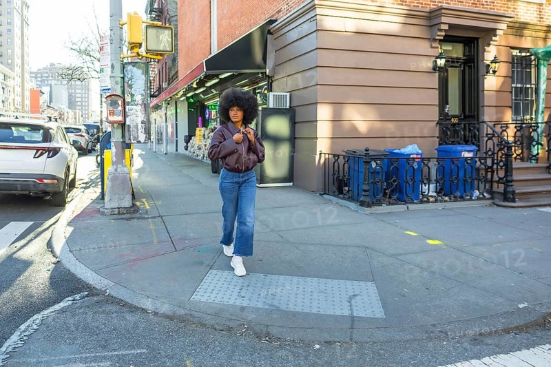 Young woman with natural afro hair walking confidently along a residential brownstone building on a sunny new york city sidewalk, showcasing urban street style in manhattan