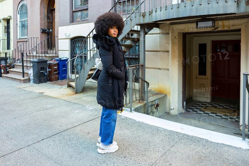 Young woman with large afro hair wearing a black winter coat and blue jeans standing on a city street sidewalk, looking attentively to the side while posing in an urban setting