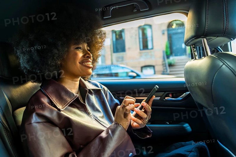 Young woman with afro hairstyle smiling, riding in the backseat of a car through new york city, using her smartphone for communication or navigation during an urban commute