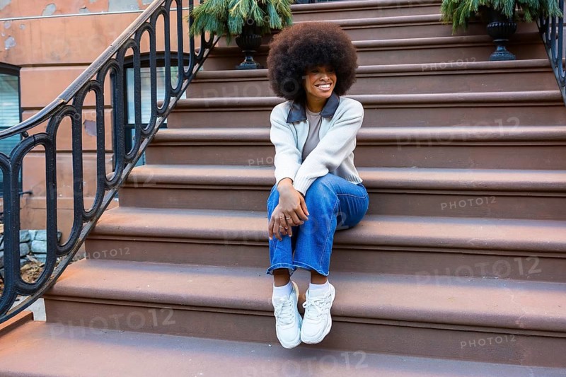 Young black woman sitting on brownstone steps in harlem, smiling and relaxed in casual street styleblue jeans, sweater and sneakers embodying urban confidence and joy