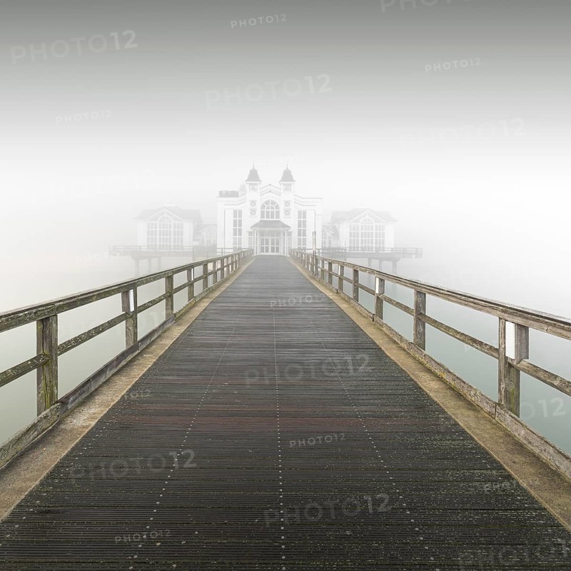 A long wooden walkway leads through thick fog to a white building, view of pier in Sellin in thick fog