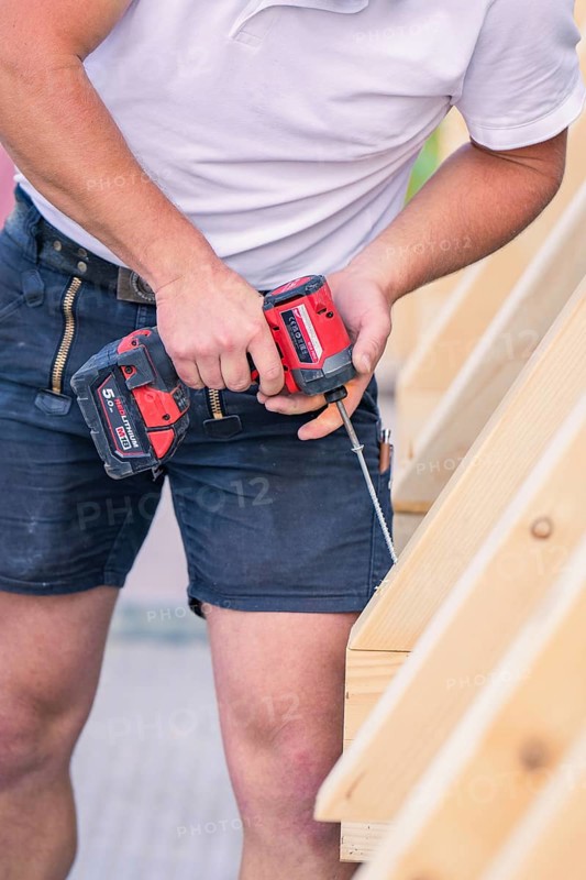A construction worker in shorts holds a screwdriver and works on a wooden structure, carpentry, new building, Stuttgart, Germany