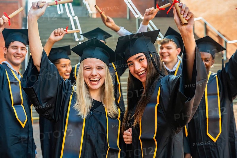 Group of happy student graduates wearing caps and gowns, cheering and holding rolled up diplomas, celebrating their academic achievement and success on campus