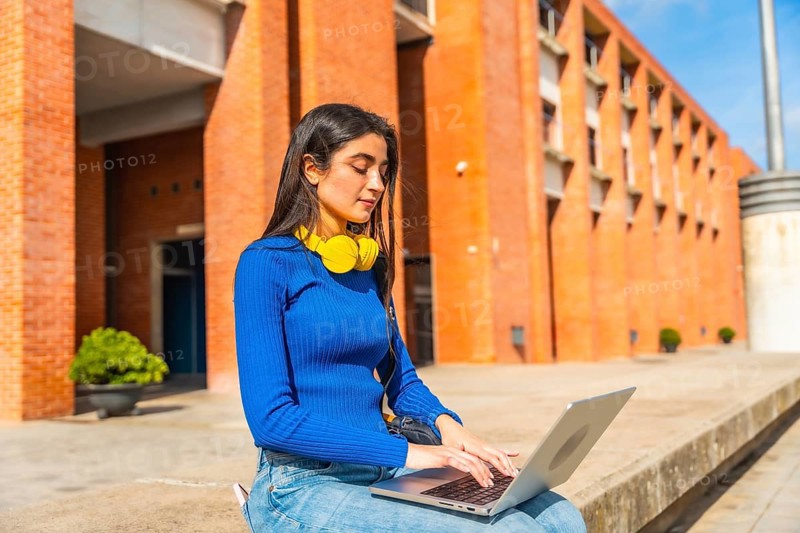 Young woman in casual clothing engaging in remote learning or project work on a laptop, sitting outside on a university campus building in daylight with headphones around her neck
