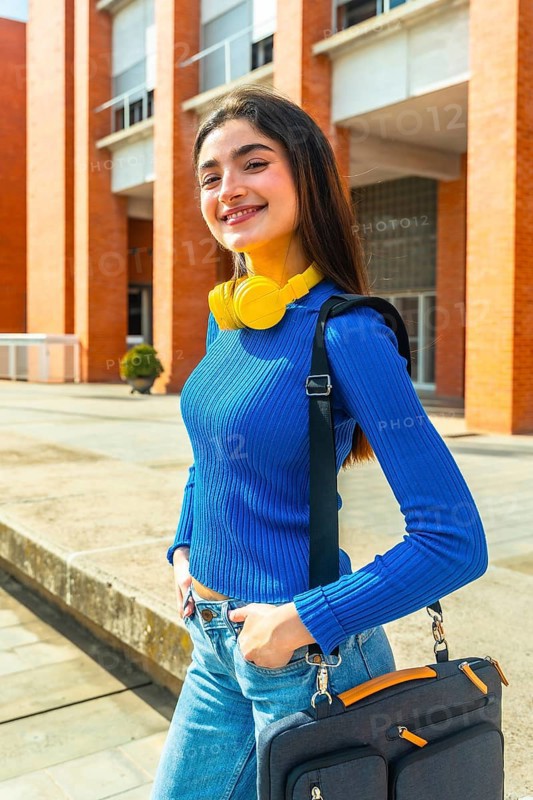 Young woman student smiling at the camera on a bright university campus, carrying a laptop bag with headphones around her neck, embodying education, youth, and learning