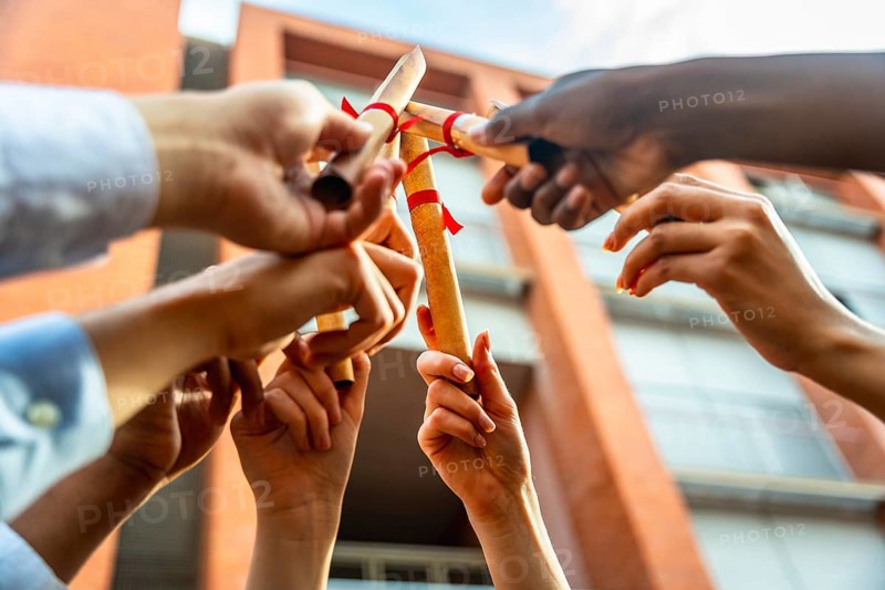 Diverse group of university students holding rolled up diplomas with red ribbons in the air against a campus building, celebrating their academic achievement and future success