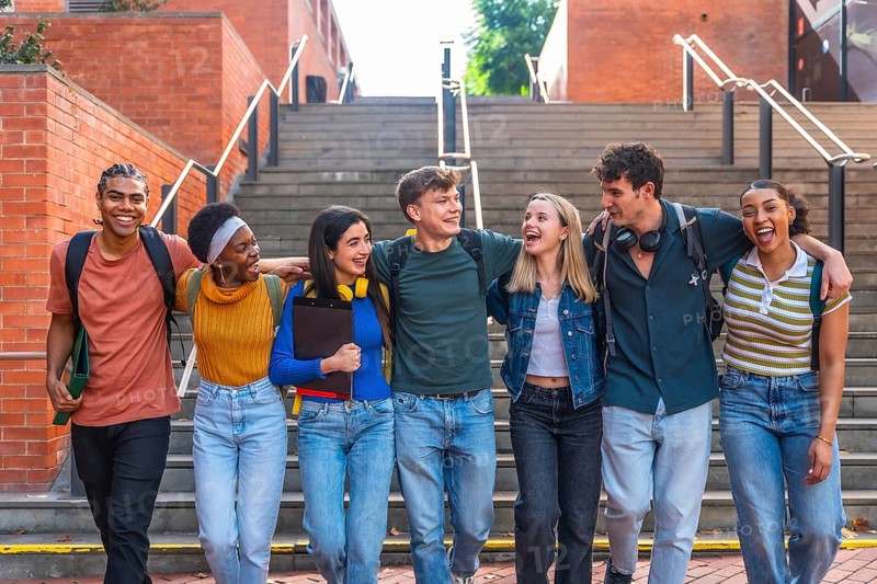 Group of happy young adult students from different ethnic backgrounds walking together on a university campus, arms around each other, laughing and enjoying their friendship