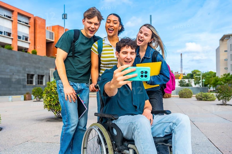 Group of happy young university students, including a man in a wheelchair, smiling and laughing while taking a selfie together outdoors on campus, celebrating friendship and inclusion