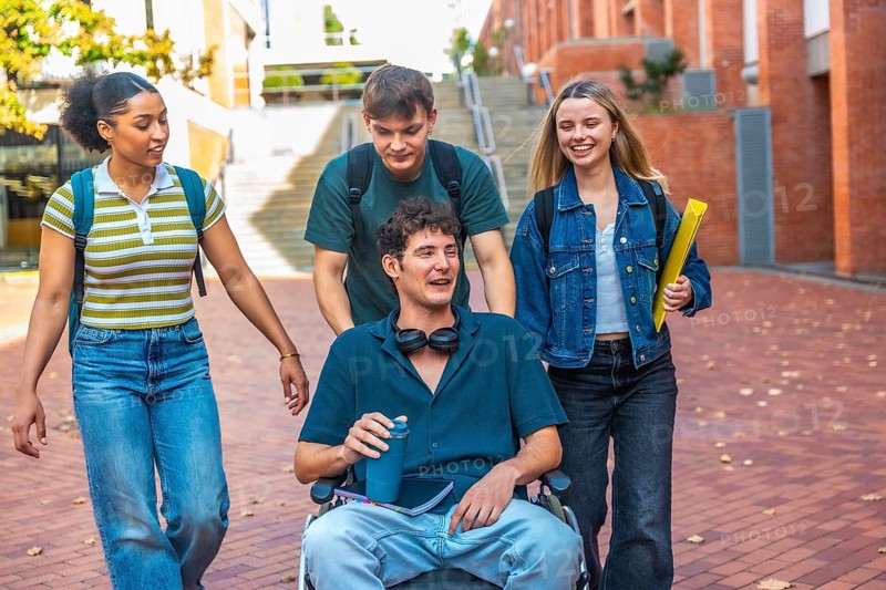 Group of diverse university students walking together on campus, one male student using a wheelchair being pushed by a friend, all engaging in conversation and smiling