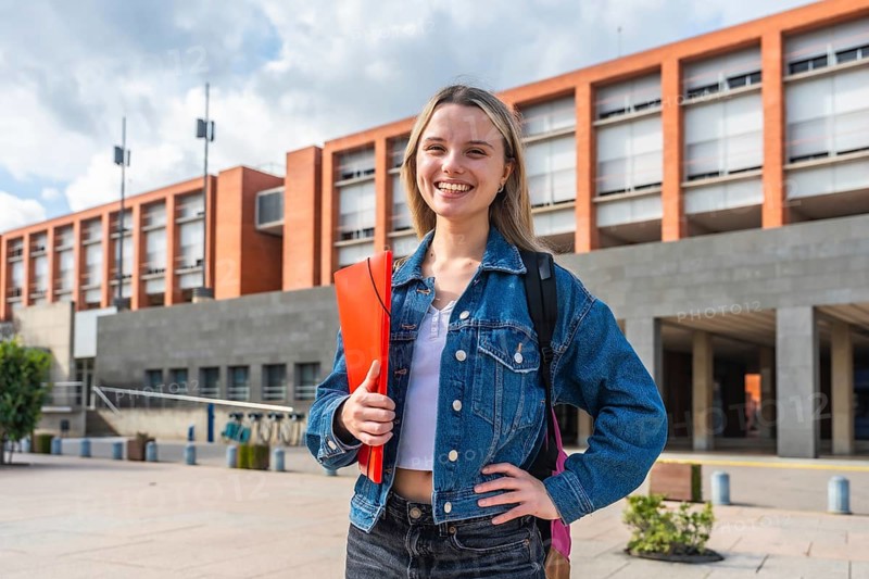Happy young female student with backpack and folder smiling, standing in front of a modern university building, representing education, future, and academic life