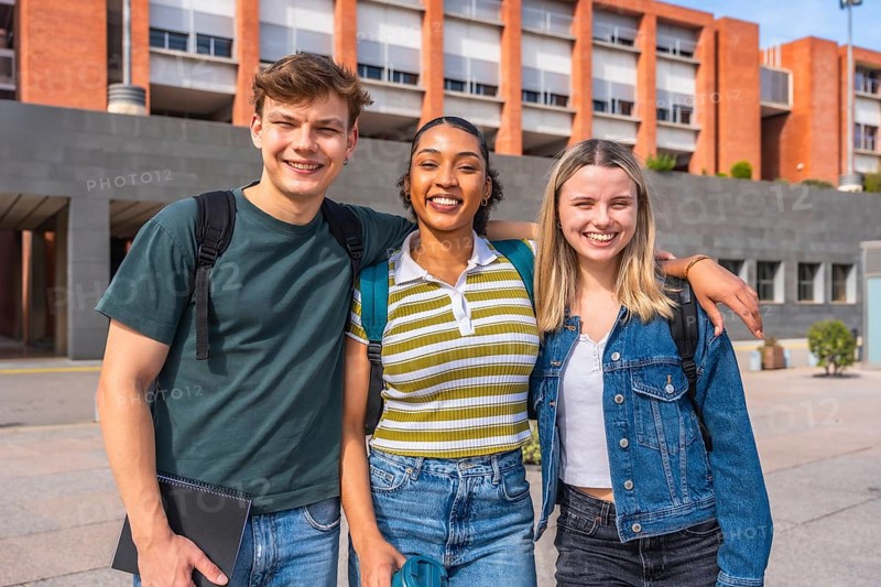 Three young multiethnic students with backpacks posing and smiling at the camera, standing in front of a modern brick university building on a sunny day