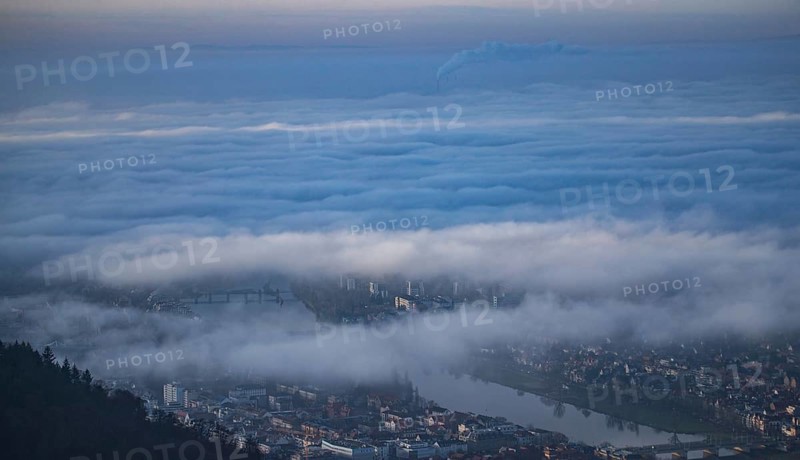 Aerial view of the city of Heidelberg on the Neckar, sea of fog from low-hanging clouds and fog fields, sunlit above the roofs of the city and the Neckar river, floating and waving with large and smoking chimney vents sticking out of the fog in the background, Heidelberg, Baden-Württemberg, Germany