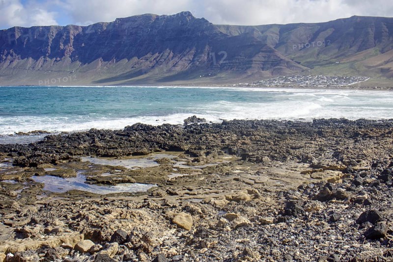 Rough coastal landscape with rocks in the foreground and mountains in the background (Risco de Famara) under a cloudy sky, Playa Caleta de Famara, Teguise Lanzarote