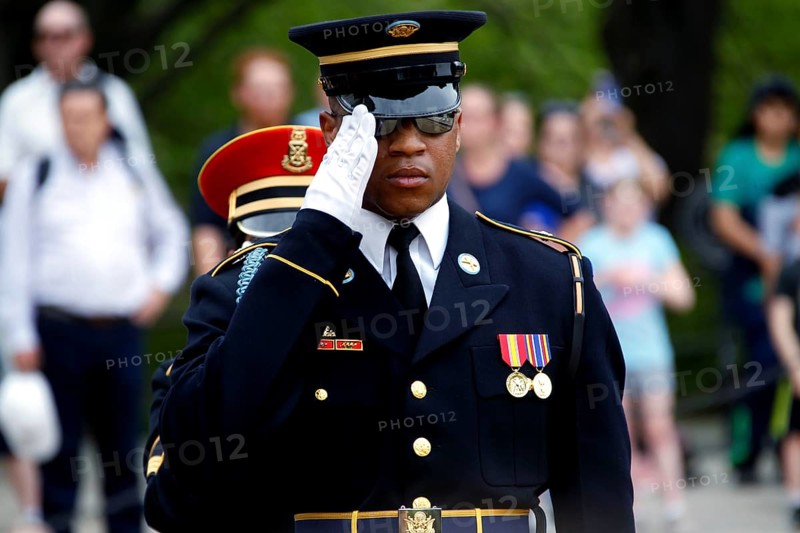 Soldier in uniform salutes during a solemn ceremony, spectator in the background, Washington D.C, USA