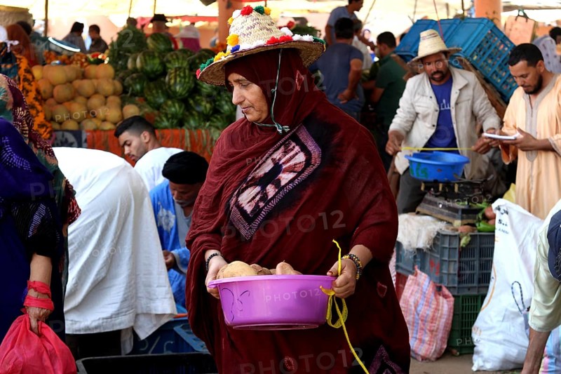 Woman wearing a headscarf carrying a basket full of vegetables at a busy market, Guelmim, Morocco