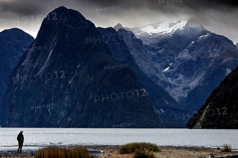 Lonely hiker on the banks of Milford Sound, with snow-capped mountains in the background, zero