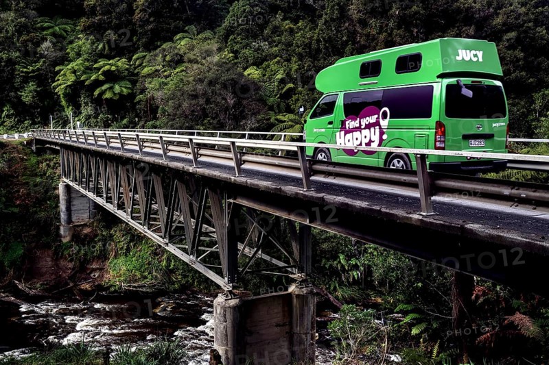 A Jucy camper crosses a bridge across the Tangarakau River on the Forgotten World Highway, surrounded by thick forest, zero