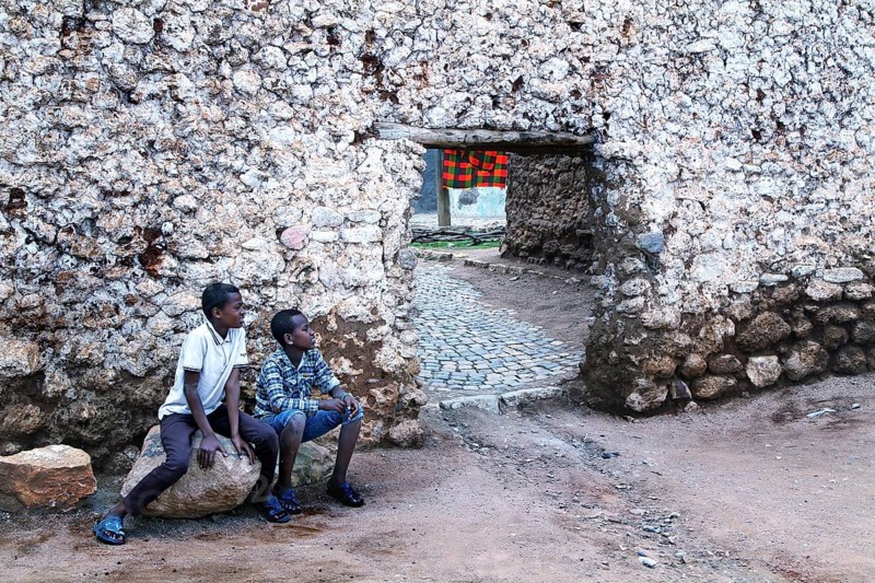 Two boys sit in front of a hyena gate in an old stone wall in the old town of Harar, Harar, Harar, Ethiopia