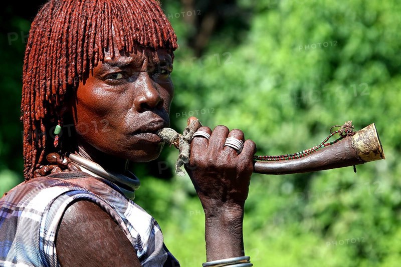 Hamer woman blowing a traditional horn surrounded by green vegetation, Turmi, Ethiopia