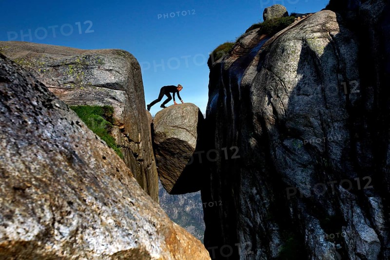 Tourist balancing on Kjeragbolten between imposing cliffs under blue sky, Lysefjord, Rogaland, Norway
