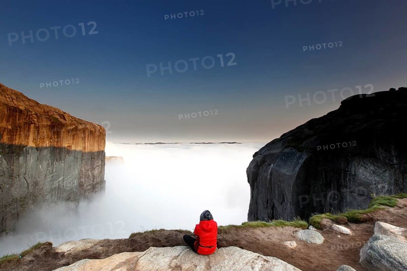 Person wearing red outfit admiring foggy view at Kjeragbolten, Lysefjord, Rogaland, Norway