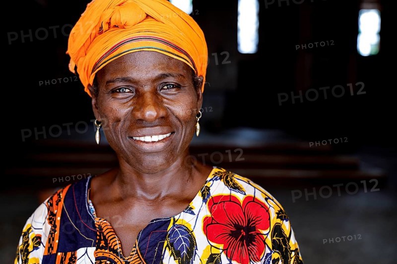 Woman in colorful dress with yellow headscarf smiles at the camera. Dark background, Bouaké, Centre, Côte d'Ivoire