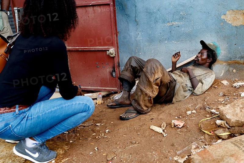 Mentally ill homeless person sits in front of a door. A woman approaches him on a dusty road, Bouaké, Côte d'Ivoire
