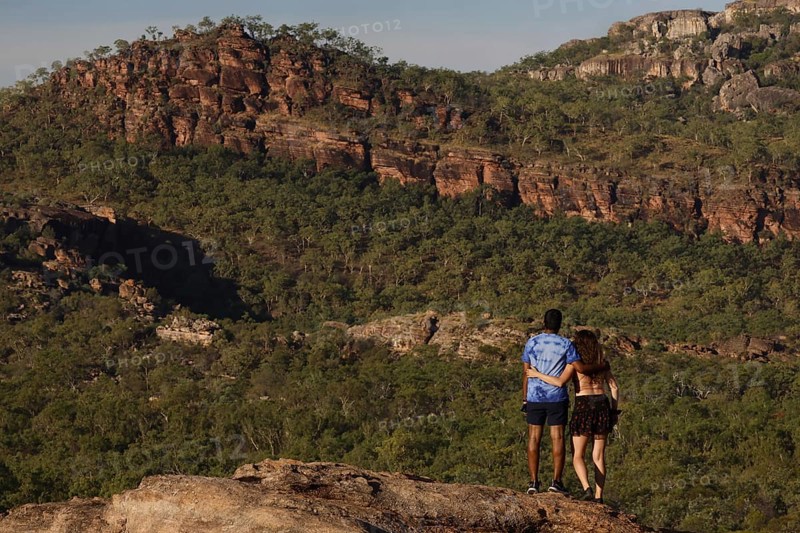 Two people on a rock with a view of wooded rocky landscape, Nawurlandia, Northern Territory, Australia
