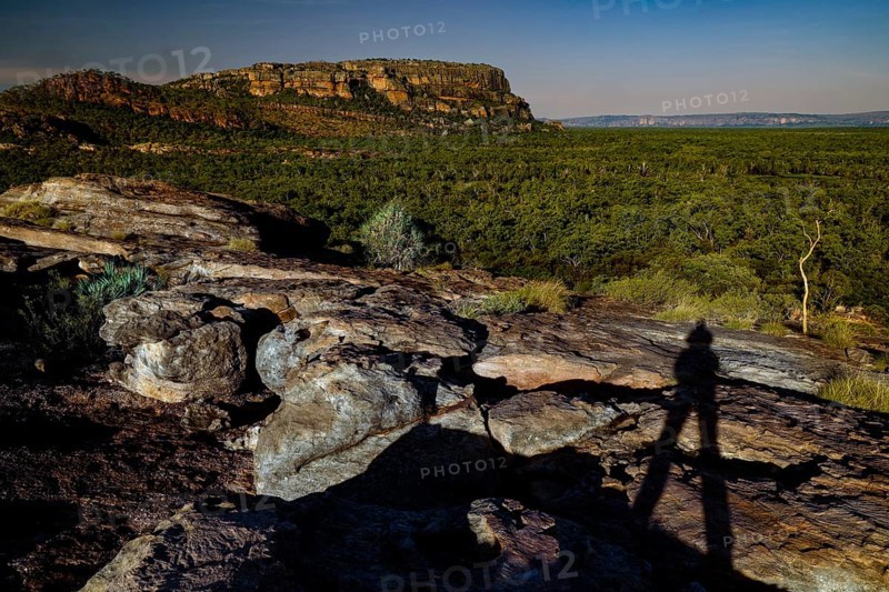 Shadows of a person on a rocky elevation in front of forest and rocks under clear sky, Nawurlandia, Northern Territory, Australia